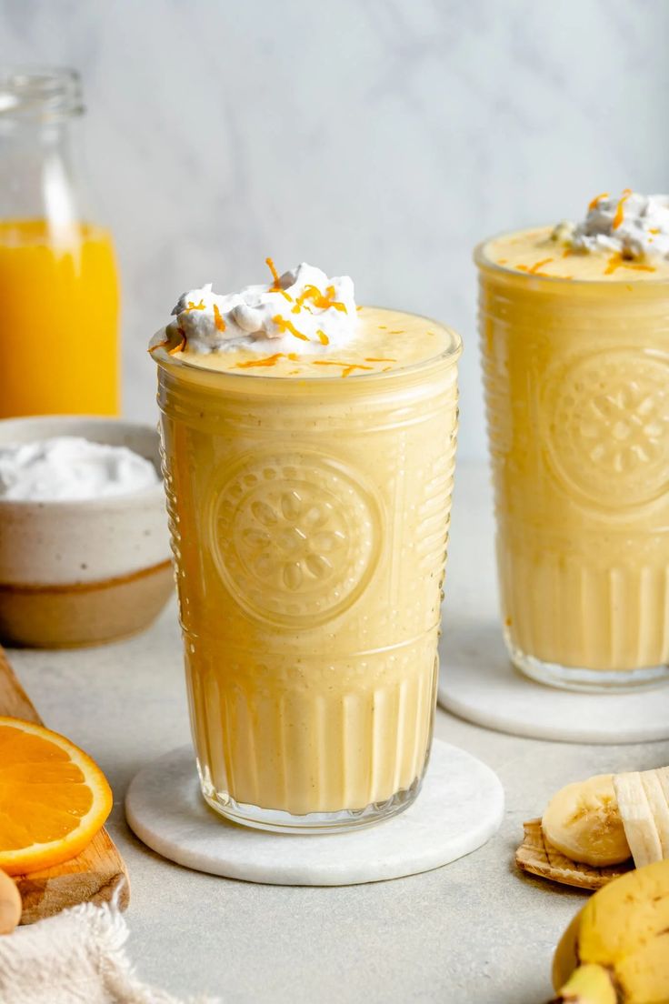 two glasses filled with orange smoothie next to sliced bananas and other fruit on the table