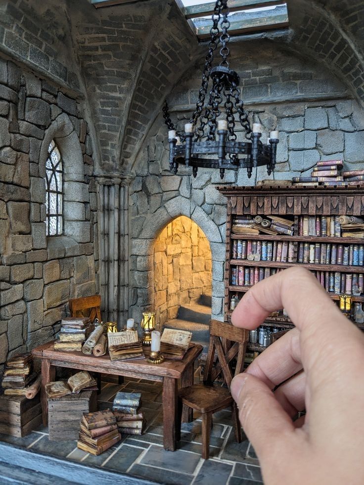 a hand is pointing at a miniature model of a room with a fireplace and bookshelf