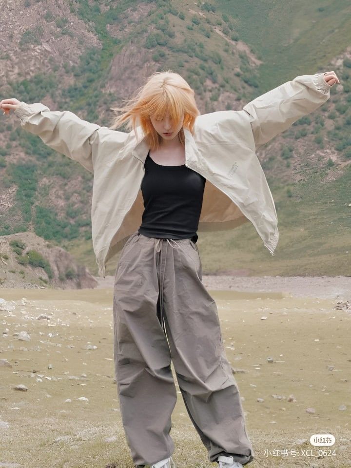 a woman with red hair standing on top of a grass covered field next to a mountain