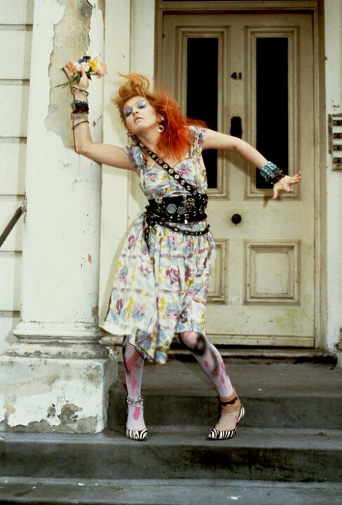 a woman with red hair and makeup is leaning against a wall holding a flower in her hand