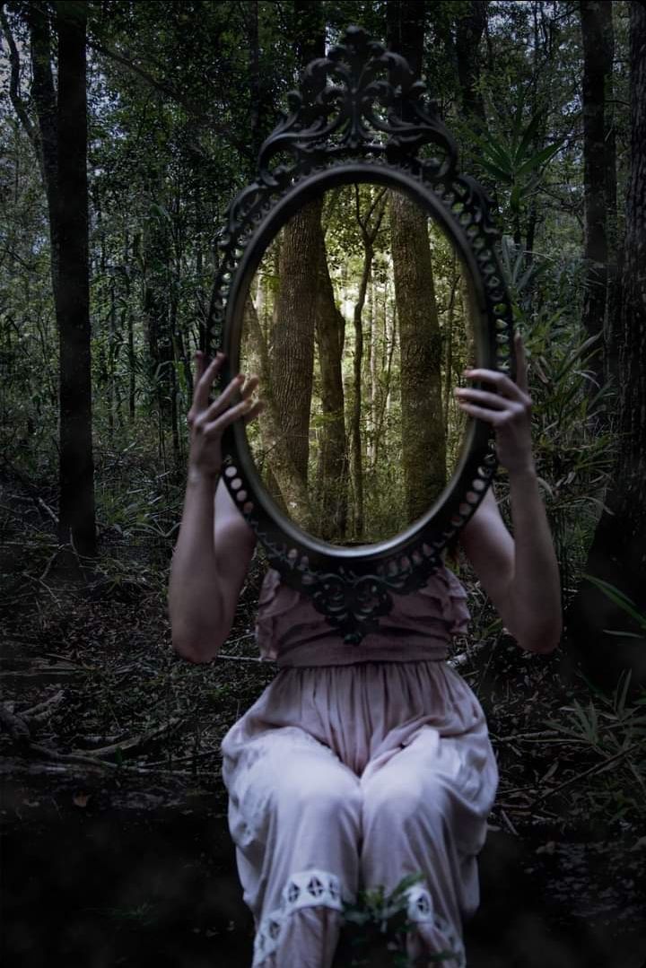 a woman sitting in the woods looking at her reflection in a mirror with both hands