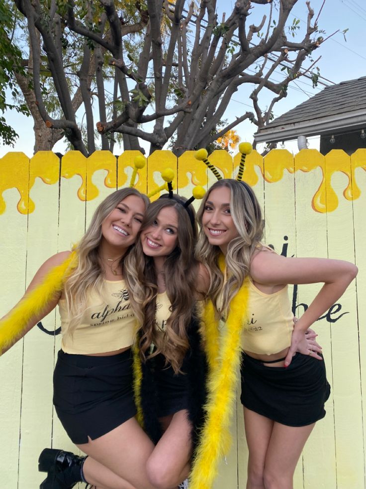 three beautiful young women standing next to each other in front of a wooden fence with yellow paint on it