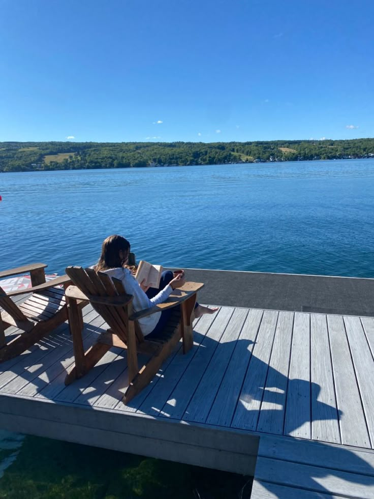 a woman is sitting on a wooden bench by the water reading a book while looking at her phone