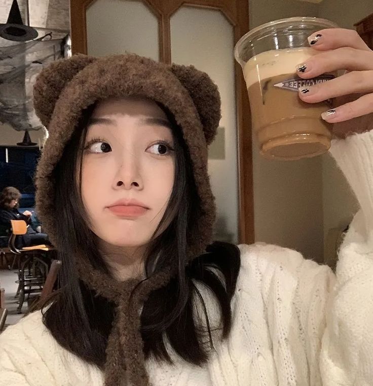 a woman wearing a bear hat and holding a drink in front of her face while looking at the camera