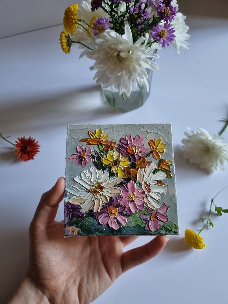 a hand holding up a card with flowers in it and a vase full of daisies