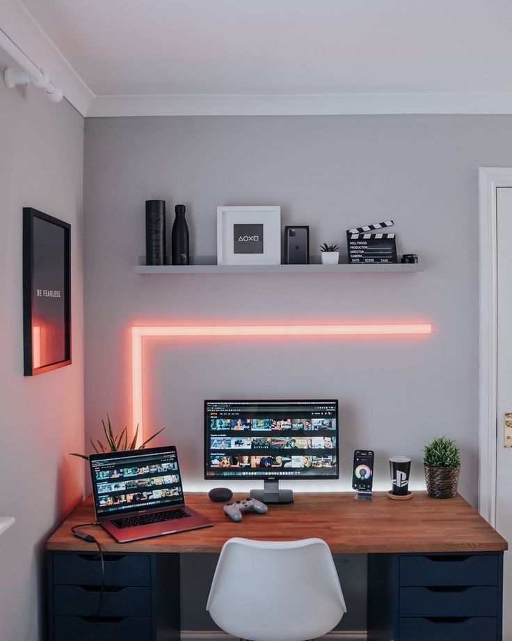 a desk with a laptop, monitor and keyboard on it in front of a neon light