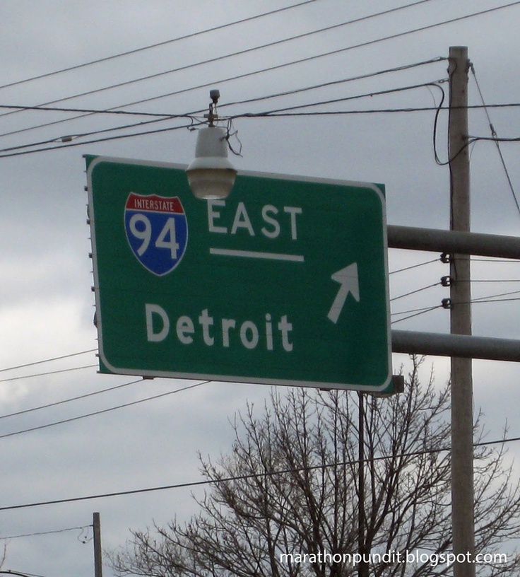 a green street sign with an arrow pointing to the east and detroit on it's side