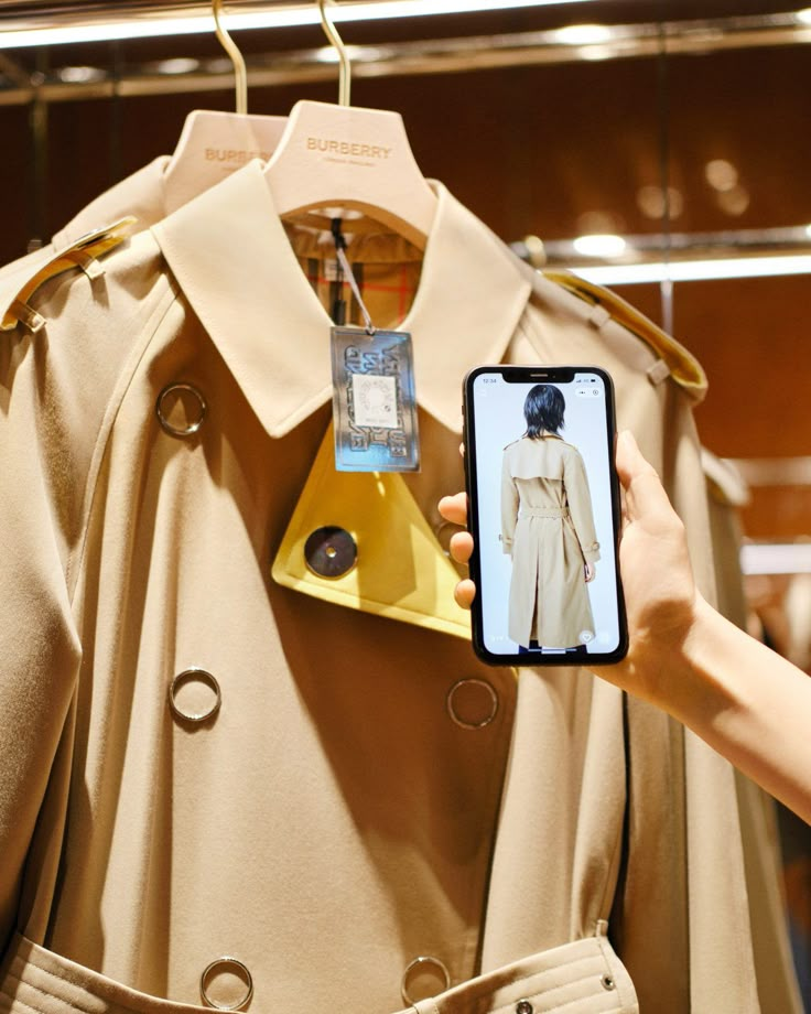 a person holding up a cell phone in front of a trench coat on a hanger