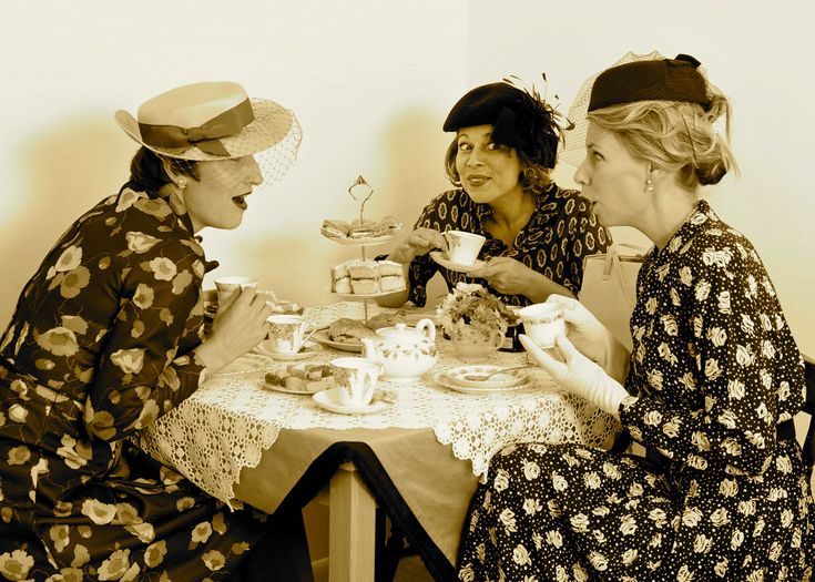 three women sitting at a table with tea cups and cake in front of their faces