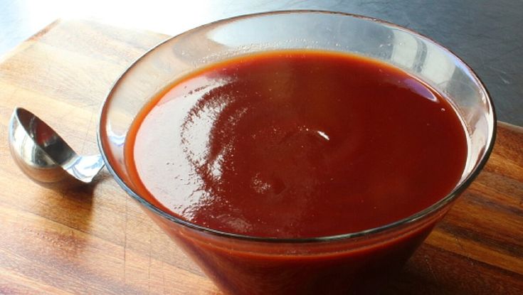 a glass bowl filled with sauce sitting on top of a wooden cutting board next to a spoon