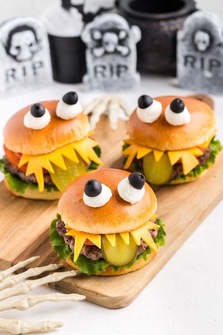 three hamburgers with fake eyes and pickles on a cutting board