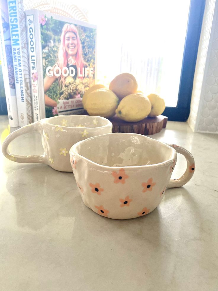 two white cups sitting on top of a counter next to lemons and a book