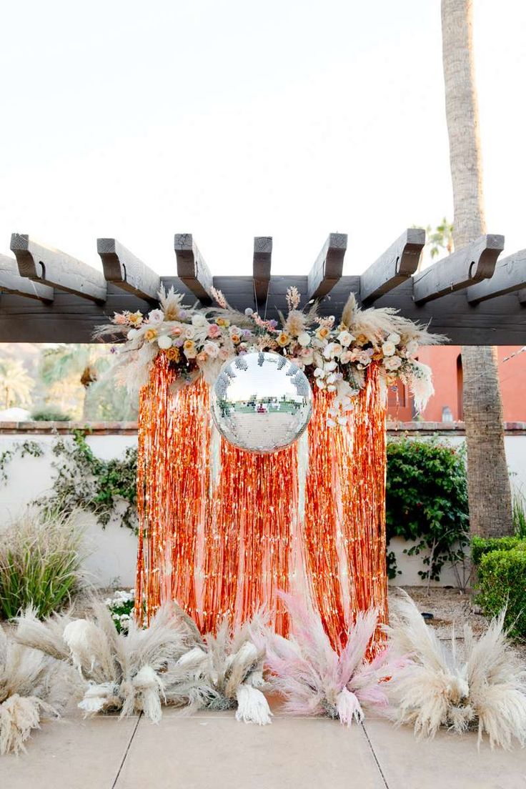 an orange and white wedding decoration with flowers, feathers and balloons on the ground in front of a wooden pergolated structure