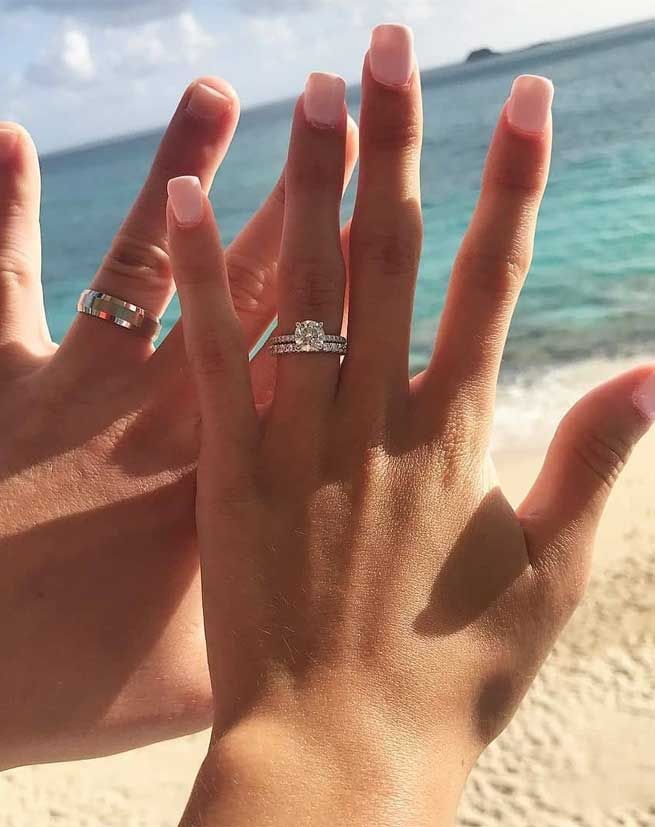 two hands with wedding rings on their fingers at the beach and water in the background