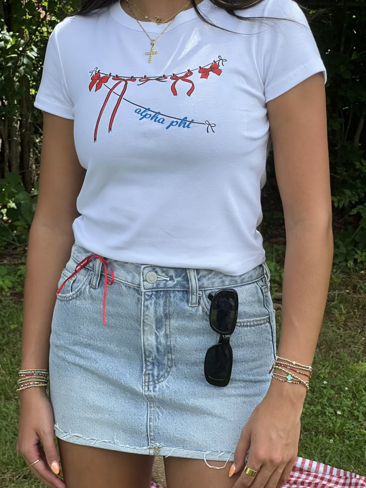 a woman standing in the grass wearing a white shirt and jean skirt with red bows on it