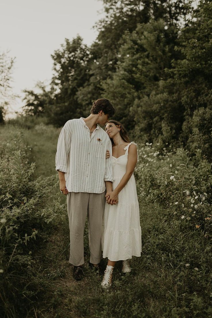 a man and woman standing next to each other in the grass with trees behind them