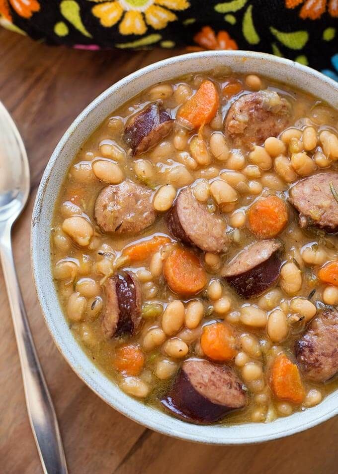 a bowl of soup with sausage, beans and carrots next to a spoon on a wooden table