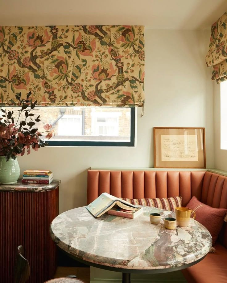 a table with books and flowers on it in front of a window that has roman shades