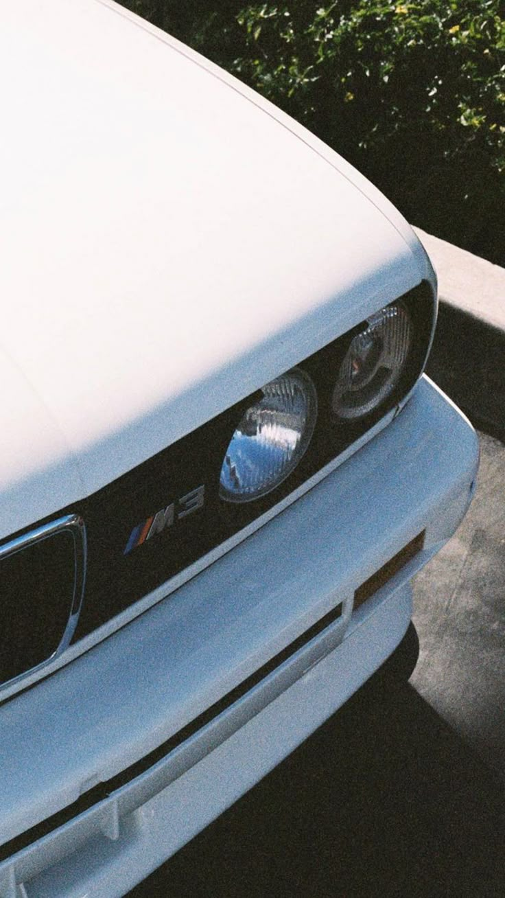 the front end of a white car parked in a parking lot next to some bushes