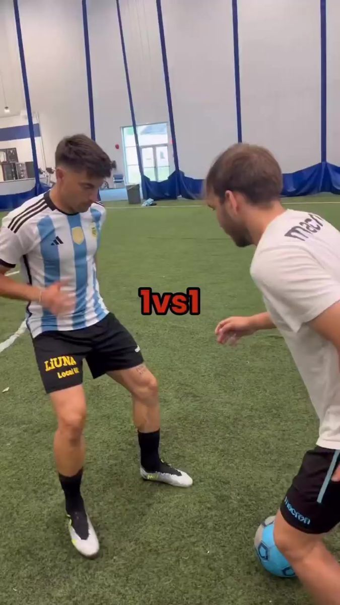 two young men are playing soccer on an indoor field, one is kicking the ball