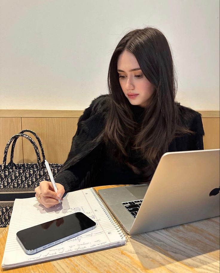 a woman sitting at a table with a laptop and notebook in front of her, writing on a notepad