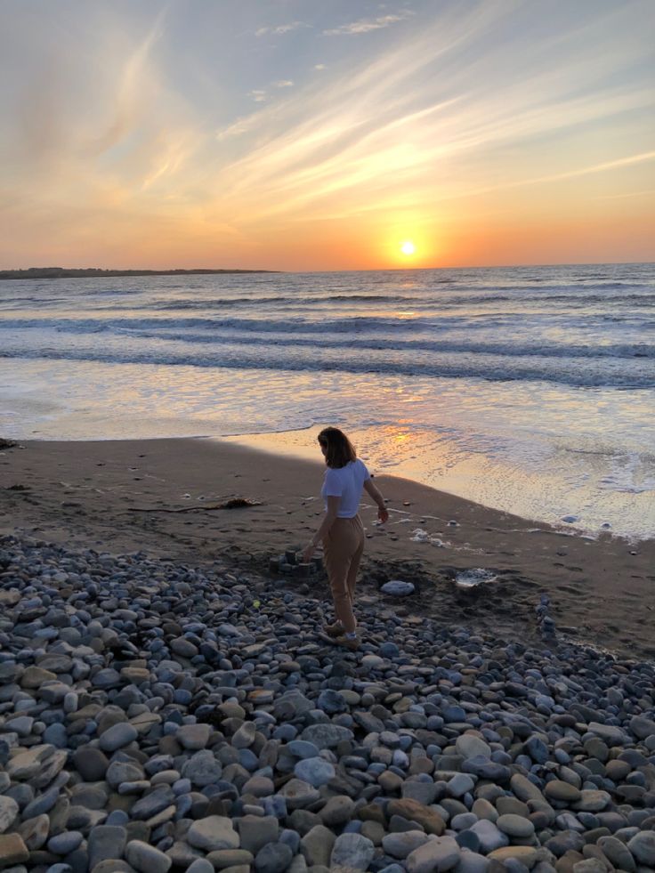 a woman standing on top of a rocky beach next to the ocean at sunset or dawn