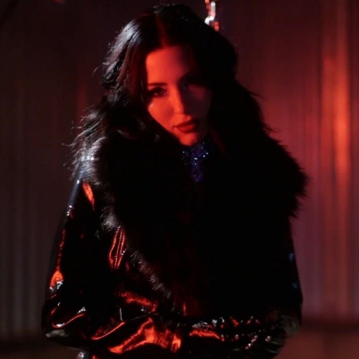 a woman with long hair wearing a black leather jacket and fur collar standing in a dimly lit room