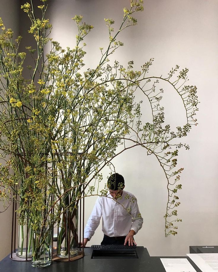 a man standing in front of a tall vase filled with flowers and greenery on top of a black table