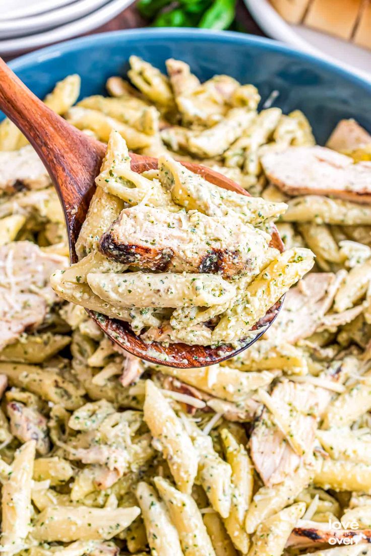 a wooden spoon full of pasta with chicken and pesto sauce on it, in a blue bowl