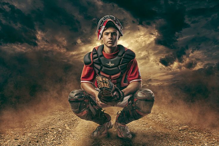 a man in catchers gear squatting down on a dirt road under a cloudy sky