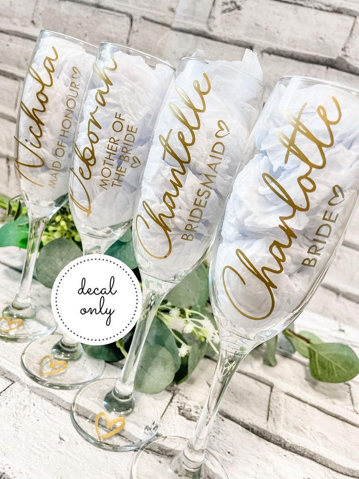 four champagne flutes with gold lettering and greenery in front of a white brick wall