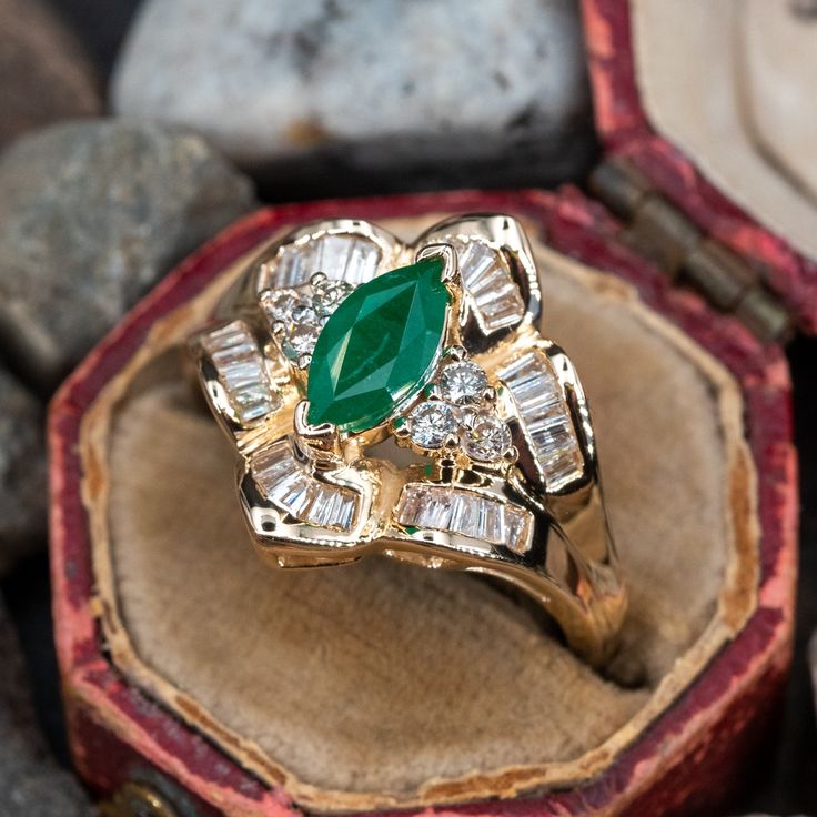 This elegant ring is centered with one (1) marquise cut natural emerald set into a two-prong setting. The emerald is bordered with six (6), prong set, round brilliant cut diamonds and thirty (30), channel set, baguette cut diamonds. The ring measures 16.0mm at the top, rises 7.8mm above the finger, tapering to 2.0mm wide and 1.1mm thick at the base of the shank. This ring is currently a size 6.75. There is a faint surface line inclusion on the emerald. Luxury Yellow Gold Emerald Ring Marquise Cut, Luxury Yellow Gold Emerald Ring With Intricate Design, Fine Jewelry Marquise Cut Emerald Ring With Diamond Accents, Green Marquise Diamond Ring Fine Jewelry, Marquise Emerald Diamond Ring In Green, Marquise Emerald Diamond Ring, Marquise Cut Emerald Ring With Diamond Accents, Green Marquise Emerald Diamond Ring, Green Marquise Emerald Rings