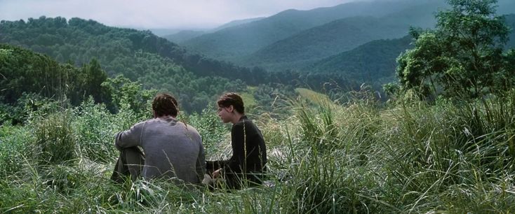 two young men sitting in tall grass looking at the mountain range with mountains in the background