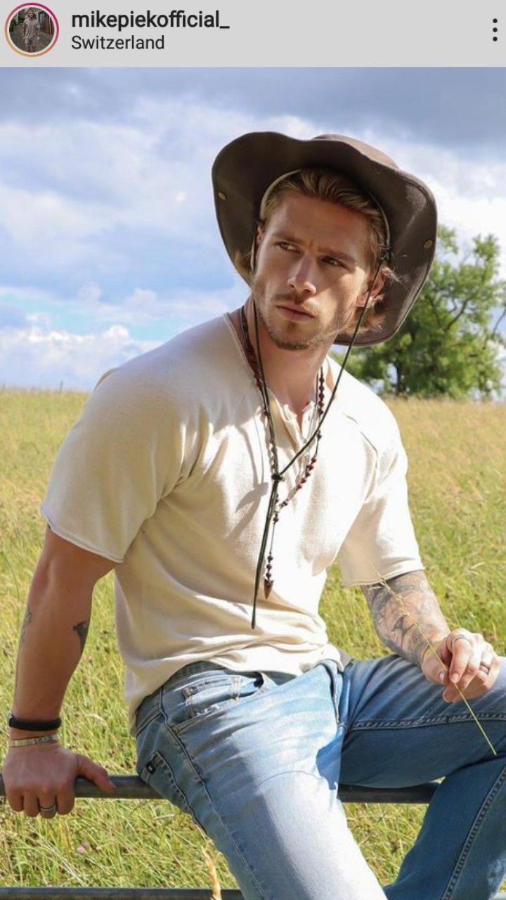 a man with a cowboy hat is sitting on a fence in the grass, wearing jeans and a t - shirt