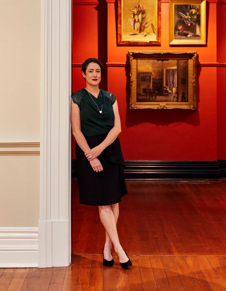 a woman standing next to a red wall with paintings on it's walls behind her