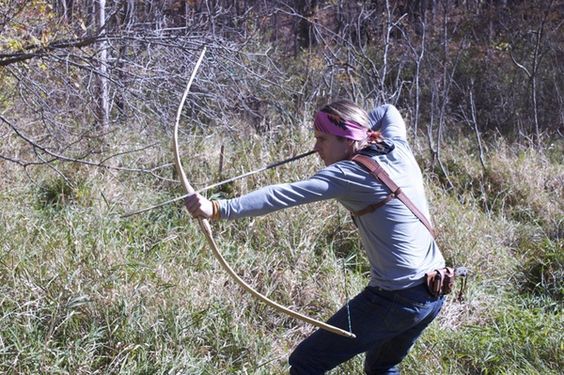 a woman is aiming an arrow with a bow in her hand while standing in the woods