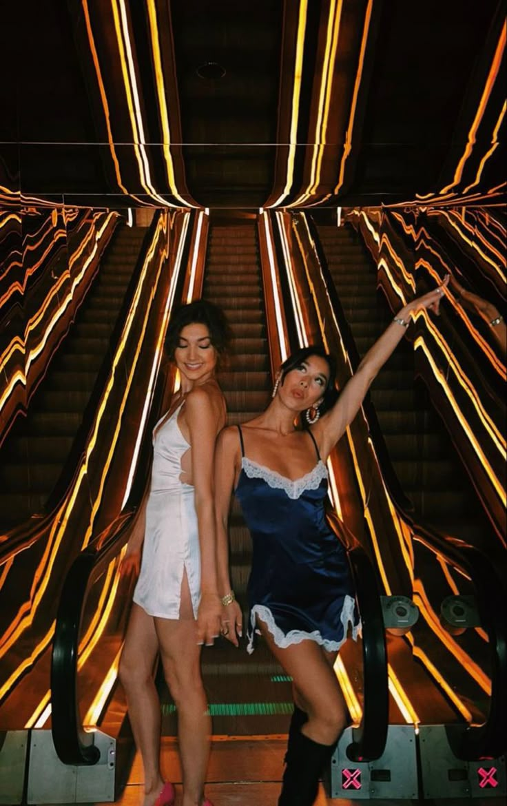 two women standing next to each other in front of an escalator with neon lights