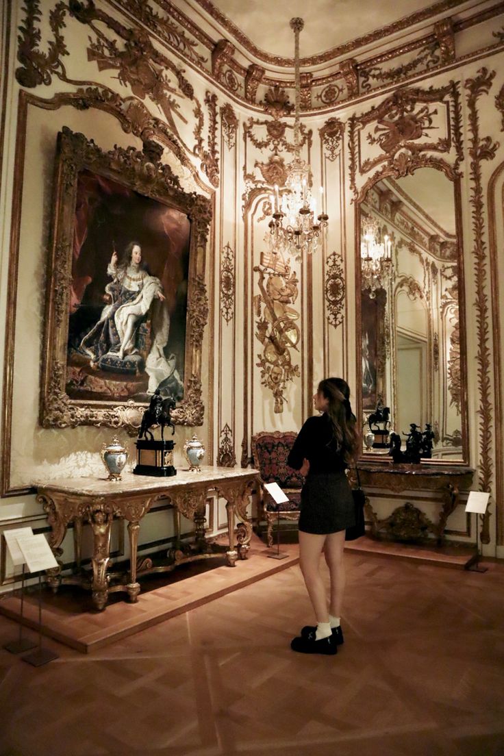 a woman standing in front of an ornate room with paintings on the walls and chandeliers