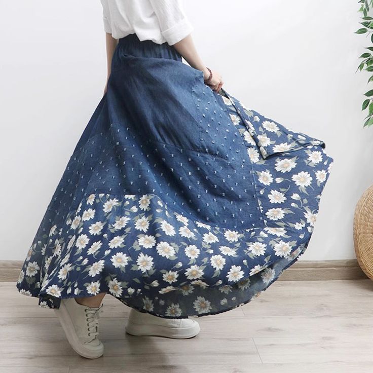 a woman in a white shirt and blue floral print skirt is standing near a plant