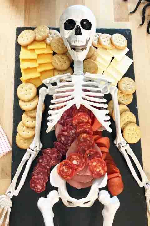 a skeleton is sitting in front of a platter of meats and cheeses