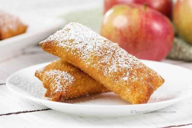 two pieces of apple turnover on a white plate with apples in the background and powdered sugar over them