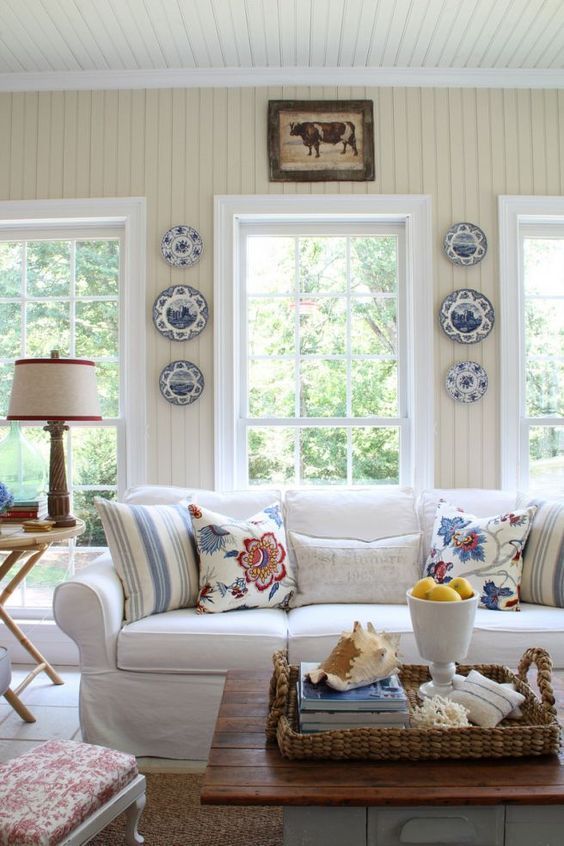 a living room filled with furniture and plates on the wall next to a table in front of two windows