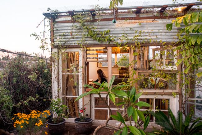 a woman is sitting in the window of a small house surrounded by plants and flowers