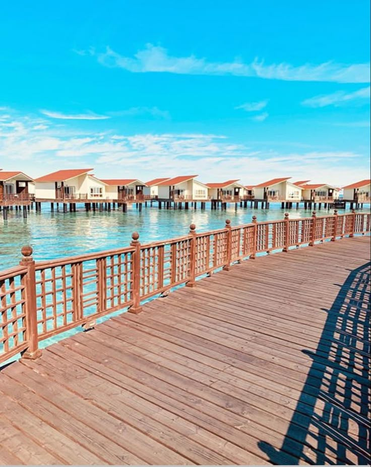 a wooden pier with houses in the background and water on both sides that is blue