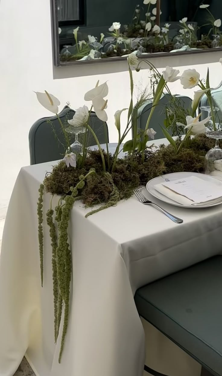 the table is set with white flowers and greenery on it, along with silverware