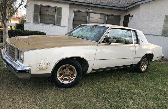 an old white car parked in front of a house with grass on the lawn and trees