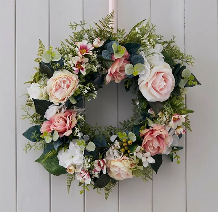 a floral wreath hanging on the side of a white wooden wall with greenery and pink flowers