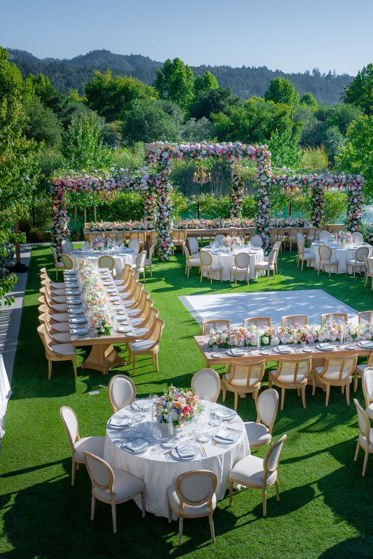 an outdoor wedding reception setup with tables and chairs set up in the grass, surrounded by greenery