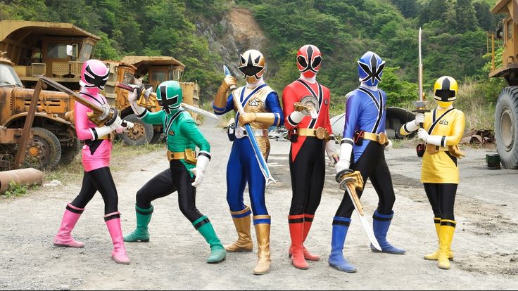 five people in power rangers costumes standing on a dirt road next to construction trucks and dump trucks