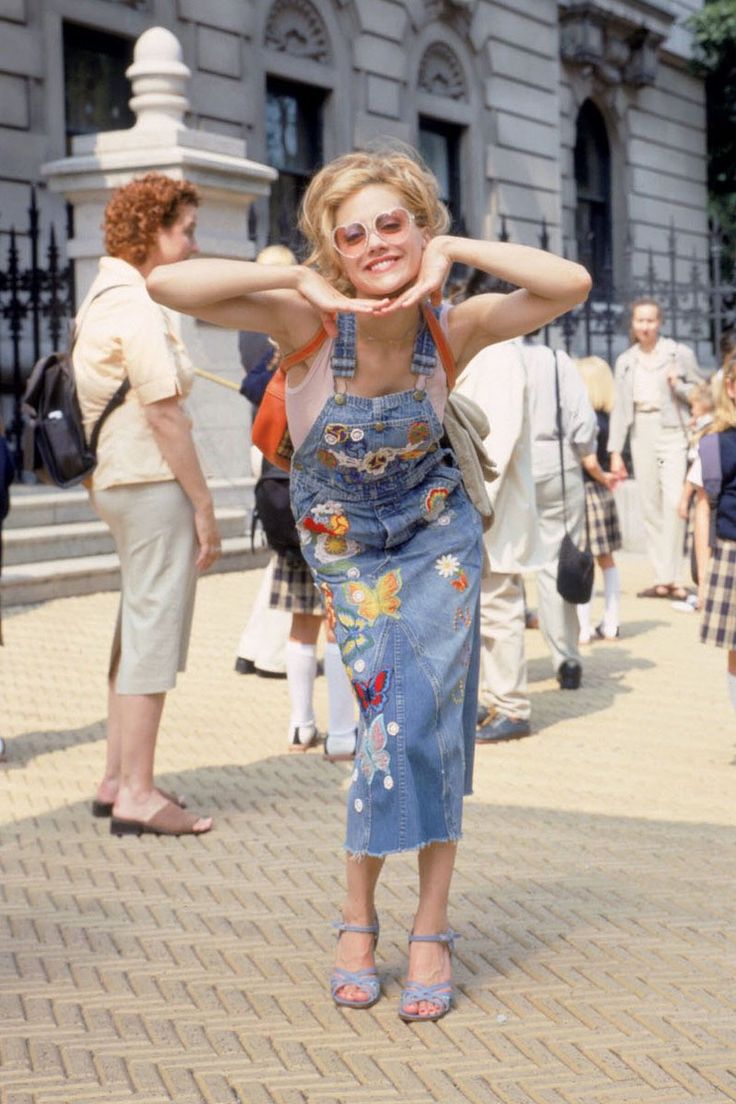 a woman in overalls standing on the sidewalk with her hands behind her head and people walking around